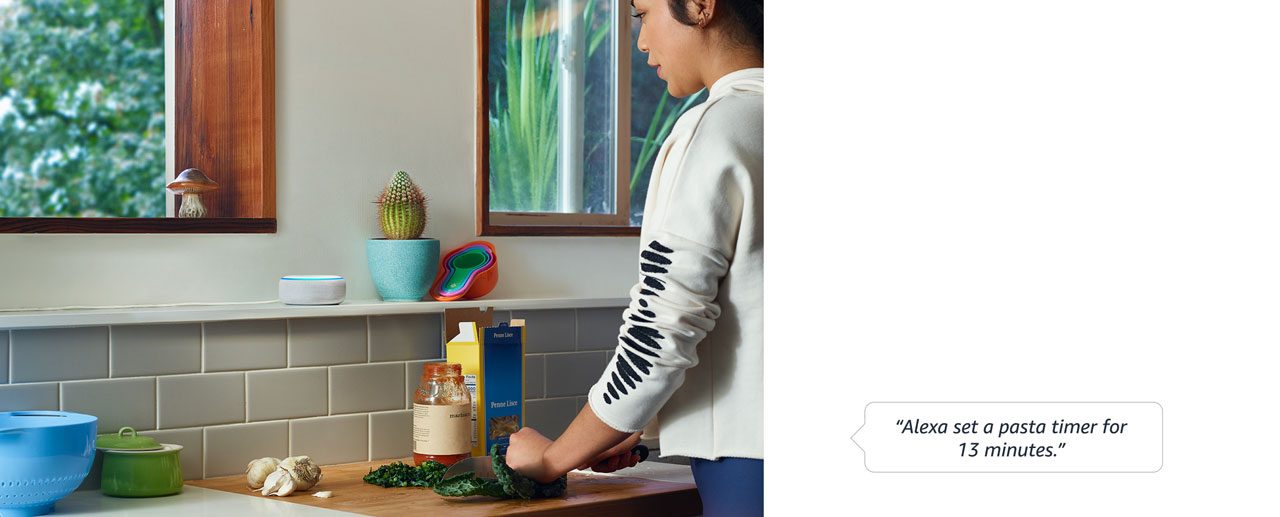 A female speaking “Alexa set a pasta timer for 13 minutes” to an Echo Dot, while processing some food materials on the kitchen counter   